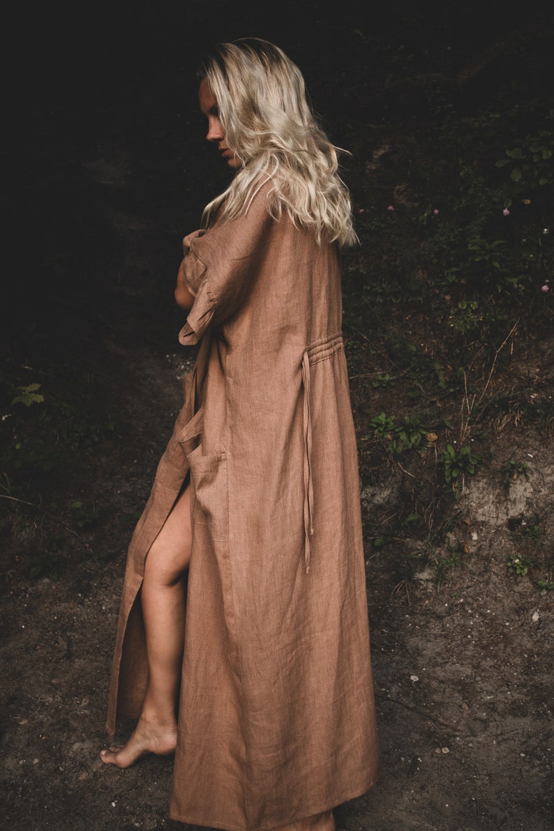 May include: A woman wearing a long, brown linen robe with a tie at the waist. The robe has a slit up the side, revealing her leg. She is standing in a forest setting.