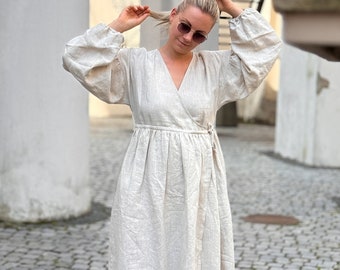 May include: A woman wearing a light beige linen wrap dress with long sleeves and a tie belt. She is wearing white sneakers and sunglasses.