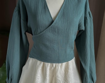 Puede incluir: Un top envolvente de color verde azulado con mangas largas y cuello en V, combinado con pantalones cortos color crema. La parte superior tiene una apariencia texturizada y los pantalones cortos tienen un dobladillo festoneado. La ropa se exhibe en un maniquí.