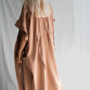 May include: A long, loose-fitting, light brown linen dress with a drawstring waist and ruffled sleeves. The dress is made of a soft, natural fabric and has a relaxed, comfortable fit.
