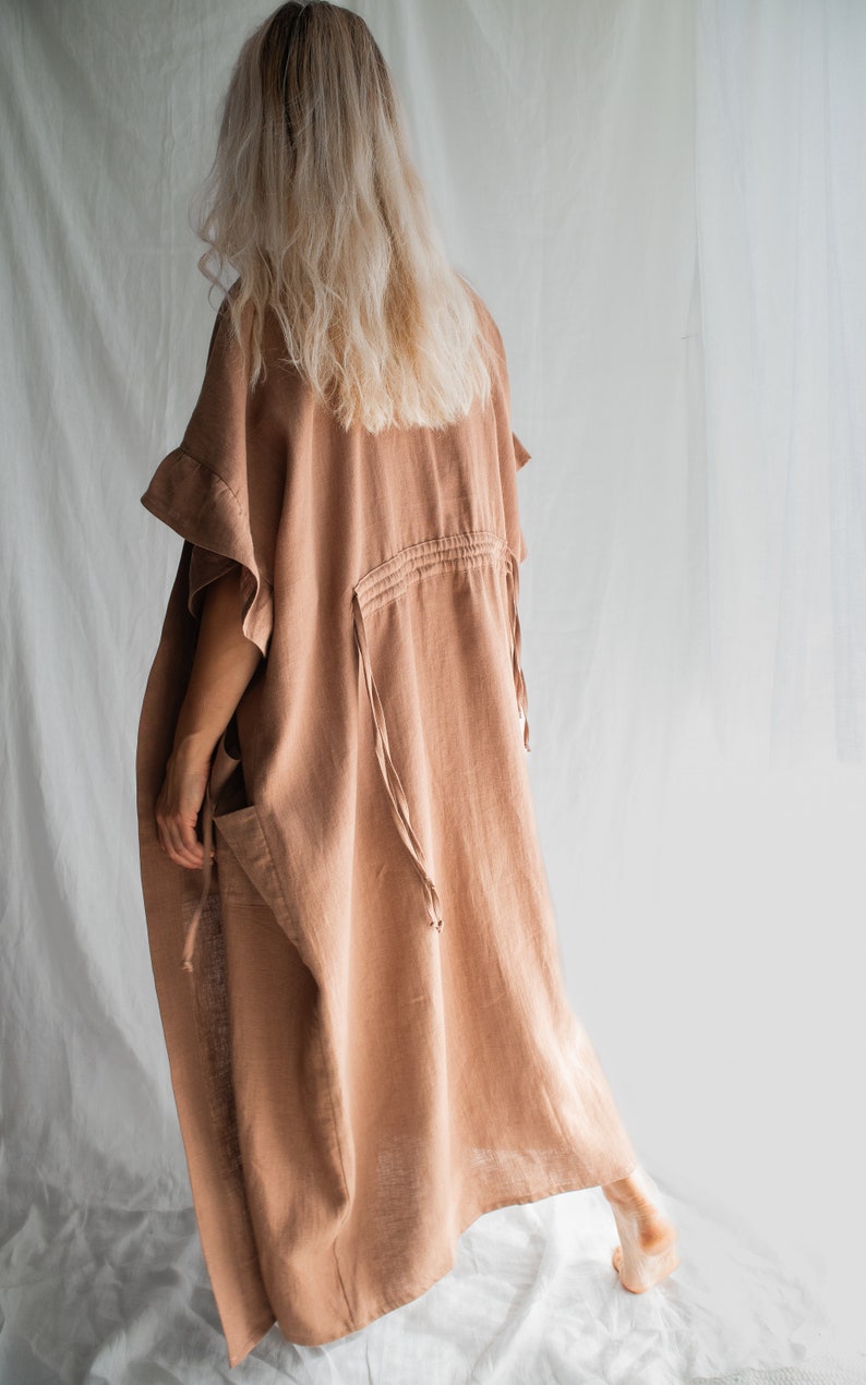 May include: A long, loose-fitting, light brown linen dress with a drawstring waist and ruffled sleeves. The dress is made of a soft, natural fabric and has a relaxed, comfortable fit.