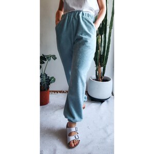 May include: A person wearing light blue sweatpants with a white top and white sandals. The person is standing in front of a potted cactus.