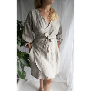 May include: A light gray linen wrap dress with long sleeves and a tie belt. The dress has a V-neckline and a relaxed fit.