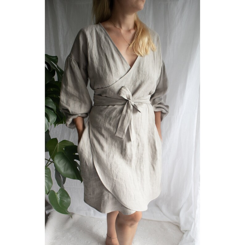 May include: A light gray linen wrap dress with long sleeves and a tie belt. The dress has a V-neckline and a relaxed fit.