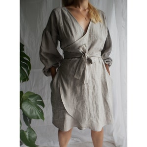 May include: A light gray linen wrap dress with long sleeves and a tie belt. The dress has a V-neckline and a flowing skirt.