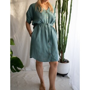 Puede incluir: Un vestido azul verdoso de manga corta con botones y cintur&oacute;n de lazo. El vestido est&aacute; hecho de una tela suave y texturizada.