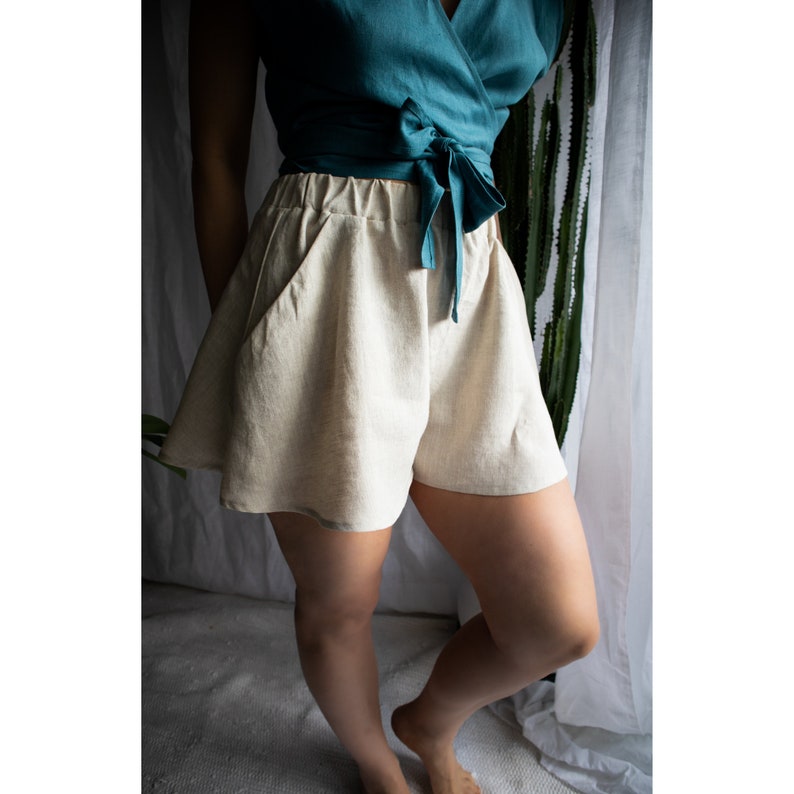 May include: High-waisted beige shorts featuring a wide-leg silhouette, an elasticized waistband, and two side pockets. The shorts are paired with a teal wrap-style top that ties at the waist with a fabric bow, creating a layered ensemble.