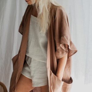 May include: A light brown linen kimono with ruffled sleeves. The kimono is open and worn over a white linen top and shorts.