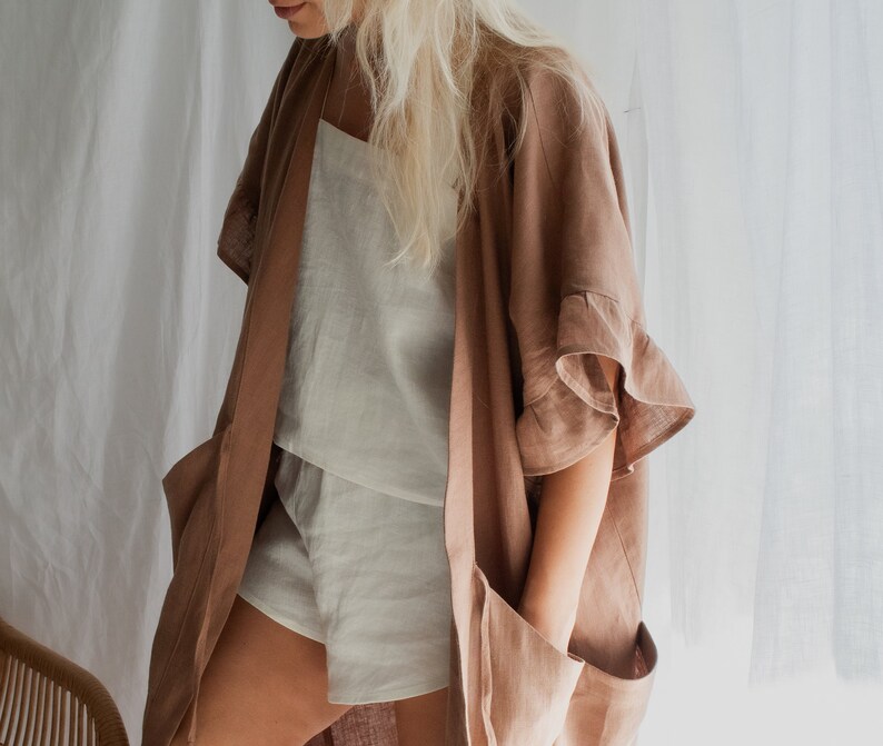 May include: A light brown linen kimono with ruffled sleeves. The kimono is open and worn over a white linen top and shorts.
