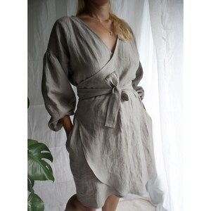 May include: A light gray linen wrap dress with long sleeves and a tie belt. The dress has a V-neckline and a relaxed fit.