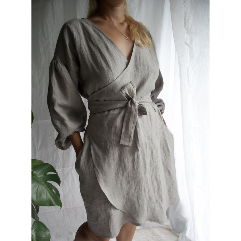 May include: A light gray linen wrap dress with long sleeves and a tie belt. The dress has a V-neckline and a relaxed fit.