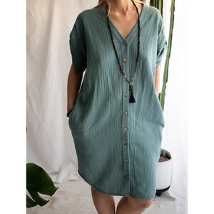 Puede incluir: Un vestido azul verdoso con botones, mangas cortas y cuello en V. El vestido tiene un corte holgado y presenta un collar con borla.