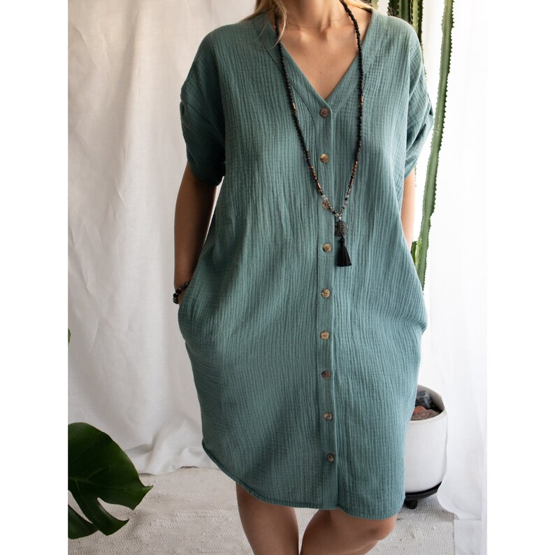 Puede incluir: Un vestido azul verdoso con botones, mangas cortas y cuello en V. El vestido tiene un corte holgado y presenta un collar con borla.