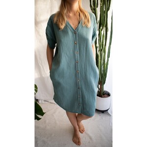 Puede incluir: Un vestido azul verdoso con botones, mangas cortas y bolsillos. El vestido est&aacute; hecho de una tela suave y texturizada.