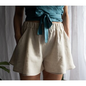 May include: Beige high-waisted shorts with a wide-leg, flared silhouette and an elasticized waistband. The shorts feature side pockets and are paired with a teal wrap-style top that ties at the front with a fabric bow at the center waist.