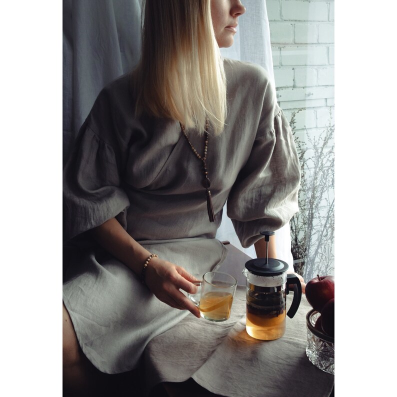 May include: A woman wearing a gray linen dress sits by a window, holding a glass of tea. A glass teapot with a black lid and handle sits on a table in front of her. The teapot is filled with tea and has a strainer inside. There are red apples in a bowl on the table.