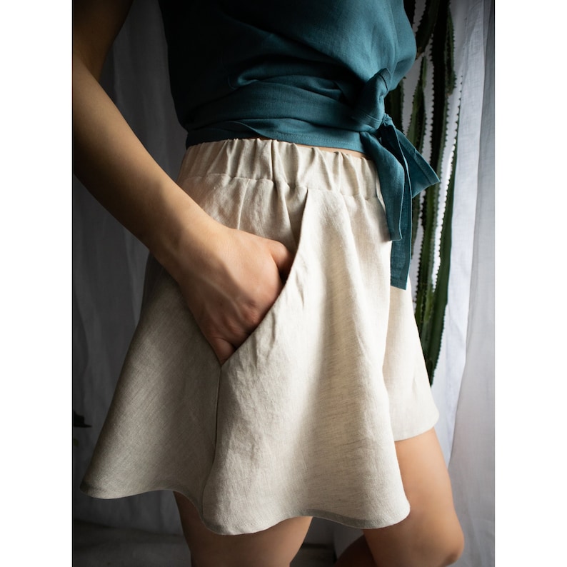 May include: A pair of beige shorts with an elastic waistband and side pockets, paired with a dark teal wrap top that features a side tie closure. The shorts have a flared, A-line silhouette and are crafted from a textured, woven fabric.