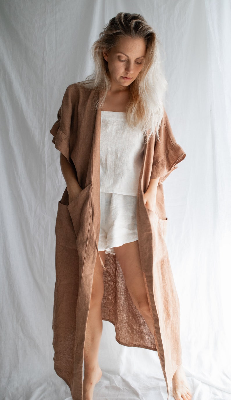 May include: A long, brown linen robe with a shawl collar and pockets. The robe is open in the front, revealing a white linen tank top and shorts underneath.