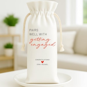 May include: A white drawstring wine bag with the text "PAIRS WELL WITH getting engaged" in black and red script. Below, it reads "CONGRATULATIONS!" with a red heart and the names "ERIN + MICHAEL". The bag has a beige rope drawstring.