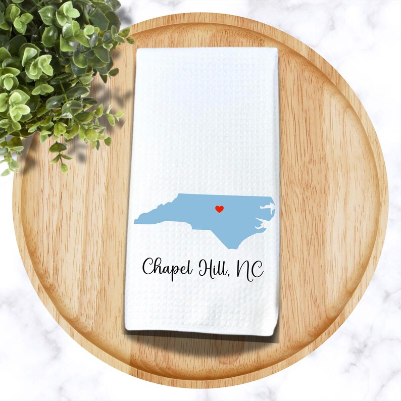 Puede incluir: Toalla de cocina blanca con un contorno azul del estado de Carolina del Norte y un coraz&oacute;n rojo sobre Chapel Hill, NC. El texto "Chapel Hill, NC" est&aacute; impreso debajo del contorno del estado.