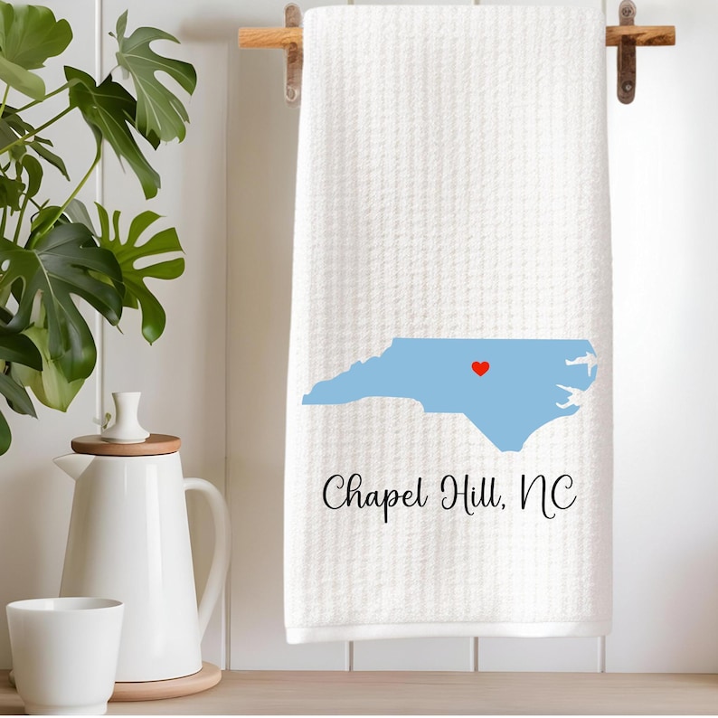 Puede incluir: Toalla de cocina blanca con un contorno azul del estado de Carolina del Norte y un coraz&oacute;n rojo sobre Chapel Hill. El texto "Chapel Hill, NC" est&aacute; impreso debajo del estado.