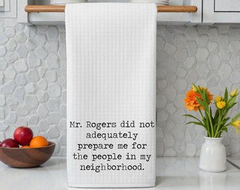 May include: A white waffle-weave kitchen towel hanging from a wooden rod. The towel features black typewriter-style text that reads: Mr. Rogers did not adequately prepare me for the people in my neighborhood. The fabric has a textured grid pattern.