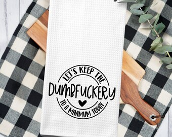 May include: A white waffle-weave kitchen towel featuring black text that reads, "LET'S KEEP THE DUMBFUCKERY TO A MINIMUM TODAY," arranged in a circular design with a small black heart icon centered at the bottom of the printed graphic.