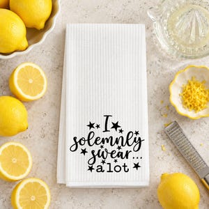 May include: A white waffle-weave kitchen towel featuring black text that reads "I solemnly swear... a lot" surrounded by small black stars. The towel is rectangular with a textured fabric surface and is centered in the frame.