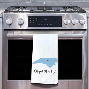 Puede incluir: Una toalla de cocina blanca con un contorno azul del estado de Carolina del Norte y un coraz&oacute;n rojo en el centro. El texto "Chapel Hill, NC" est&aacute; impreso debajo del contorno del estado.