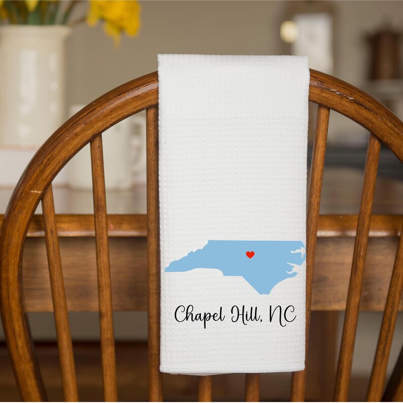 Puede incluir: Toalla de cocina blanca con un contorno azul del estado de Carolina del Norte y un coraz&oacute;n rojo sobre Chapel Hill. El texto "Chapel Hill, NC" est&aacute; impreso debajo del estado.