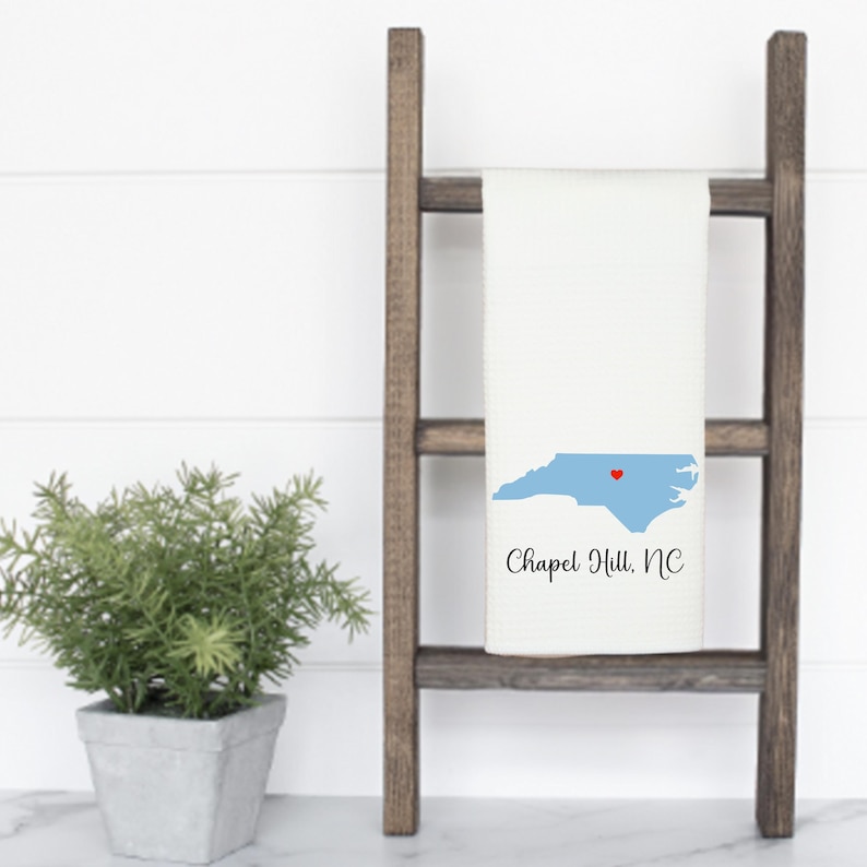 Puede incluir: Una toalla de cocina blanca con un contorno azul del estado de Carolina del Norte y un coraz&oacute;n rojo sobre Chapel Hill. El texto "Chapel Hill, NC" est&aacute; impreso debajo del contorno del estado. La toalla est&aacute; colgada en una escalera de madera.