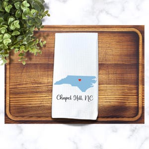 Puede incluir: Toalla de cocina blanca con un contorno azul del estado de Carolina del Norte y un coraz&oacute;n rojo sobre Chapel Hill. El texto "Chapel Hill, NC" est&aacute; impreso debajo del estado.