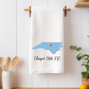 Puede incluir: Toalla de cocina blanca con un contorno azul del estado de Carolina del Norte y un coraz&oacute;n rojo sobre Chapel Hill. El texto "Chapel Hill, NC" est&aacute; impreso debajo del contorno del estado.
