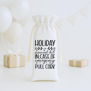 May include: A white drawstring bag with the text "HOLIDAY survival kit IN CASE OF emergency. PULL CORK" in black. The bag has a beige drawstring and is surrounded by white balloons and small wrapped gifts.