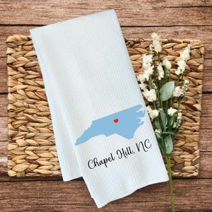 Puede incluir: Toalla de cocina blanca con un contorno azul del estado de Carolina del Norte y un coraz&oacute;n rojo en el centro. El texto "Chapel Hill, NC" est&aacute; impreso debajo del contorno del estado.