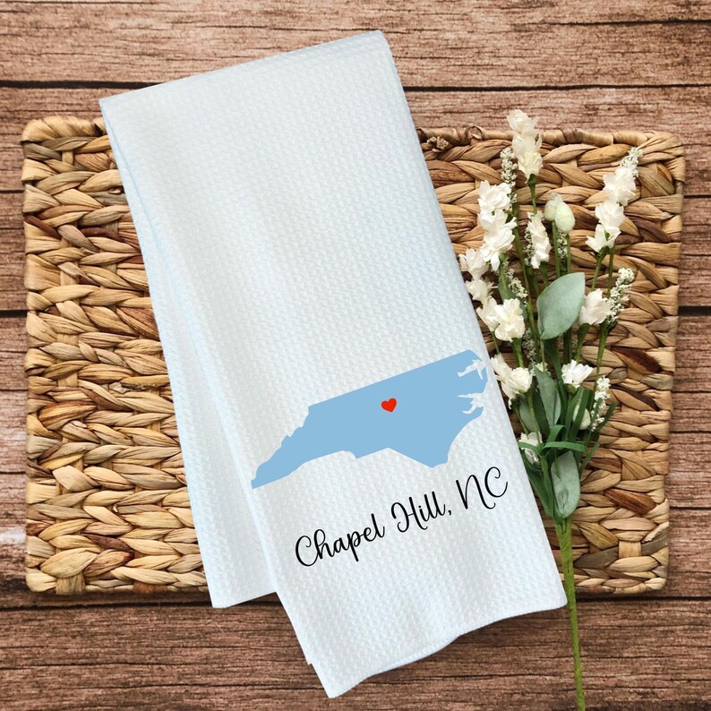 Puede incluir: Toalla de cocina blanca con un contorno azul del estado de Carolina del Norte y un coraz&oacute;n rojo en el centro. El texto "Chapel Hill, NC" est&aacute; impreso debajo del contorno del estado.