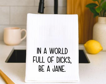 May include: A white ribbed kitchen towel featuring the black printed text: IN A WORLD FULL OF DICKS, BE A JANE. The towel has a textured surface and is displayed in a vertical orientation, showcasing the bold, sans-serif lettering clearly.