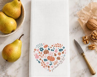 May include: A white waffle-weave kitchen towel featuring a heart-shaped graphic composed of autumn-themed illustrations including pumpkins, leaves, and flowers, with the text "give thanks to the Lord" printed along the side of the heart design.
