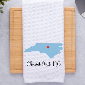 Puede incluir: Toalla de cocina blanca con un contorno azul del estado de Carolina del Norte y un coraz&oacute;n rojo sobre Chapel Hill, NC. El texto "Chapel Hill, NC" est&aacute; impreso debajo del contorno del estado.