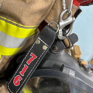 May include: Black leather keychain with the number "911" in red, white outline, and white stitching. The keychain is attached to a silver carabiner clip. The background shows a firefighter's gear, including a helmet and uniform.