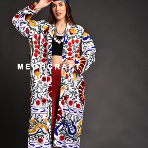 Può includere: Una giacca bianca a maniche lunghe ricamata con un motivo floreale in rosso, blu, giallo e verde. La giacca è aperta e la modella la indossa sopra un top nero e pantaloni rossi.
