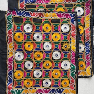 Può includere: Due quadrati tessili neri ricamati. Ogni quadrato presenta un motivo a griglia centrale di quadrati colorati con disegni circolari gialli, bianchi e blu. I bordi sono adornati da ricami multicolori intricati, tra cui rosso, verde e rosa.