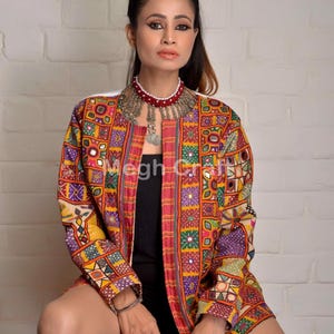 May include: A colorful embroidered jacket with mirror work and a black top underneath. The jacket has a patchwork design with various colors and patterns, including red, yellow, green, blue, and purple. The model is wearing a silver and red beaded necklace.