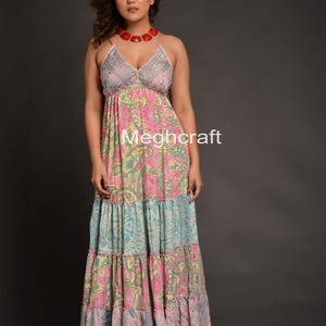 May include: A long, tiered maxi dress with a V-neck and spaghetti straps. The dress features a patchwork design with pink, blue, and green paisley patterns. The text "Meghcraft" is visible on the dress. A red necklace completes the look.