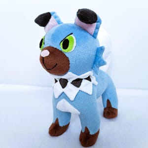 May include: A plush toy creature in shades of blue, brown, and white. It has green eyes, black ears, and a white and black collar. The toy is standing on a white surface.