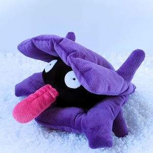 May include: A plush toy of a Pokémon character, featuring a black body, large purple petals, and a pink tongue. The toy has white eyes and is set against a snowy background.