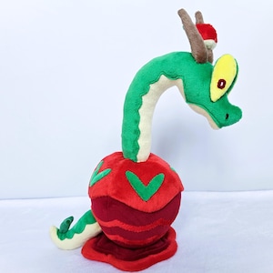 Puede incluir: Un dragón de peluche con cabeza y cuello verdes, cuerpo rojo y cola roja y verde. El dragón tiene ojos amarillos y cuernos marrones. El juguete está hecho de tela suave y tiene aspecto de personaje de dibujos animados.