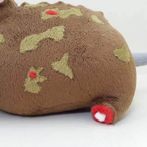 K&ouml;nnte beinhalten: Braunes Pl&uuml;schtier mit einem grauen Schwanz und gr&uuml;nen und roten Stickdetails. Das Spielzeug hat die Form einer Ratte oder Maus.