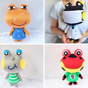 May include: Four plush animal toys, each a different frog character. The first frog is orange with brown spots and wears a blue dress. The second frog is black with yellow eyes and wears a white lab coat and a white face mask. The third frog is gray with yellow eyes and wears a green and white striped shirt. The fourth frog is red with black spots and wears a green shirt with a yellow number 6.