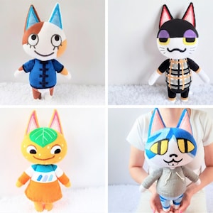 Op de afbeelding: Vier pluche katten speelgoed met verschillende ontwerpen. De eerste kat is wit met bruine vlekken en blauwe oren, draagt een blauw shirt met een mandarijnkraag. De tweede kat is zwart-wit met paarse accenten, draagt een geruit shirt. De derde kat is oranje met een groen blad op zijn hoofd, draagt een blauw en oranje shirt. De vierde kat is blauw met gele ogen, draagt een grijze hoodie.
